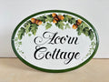 An oval decorative house name sign with a white background, dark green border, and a hand-painted wreath of oak leaves and acorns above the words 'Acorn Cottage' in black script.