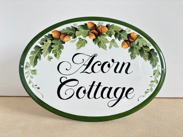 An oval decorative house name sign with a white background, dark green border, and a hand-painted wreath of oak leaves and acorns above the words 'Acorn Cottage' in black script.