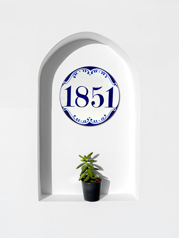 A decorative ceramic wall Address plaque featuring the house number '1851' in blue within a blue ornamental border, mounted on a white arched niche wall