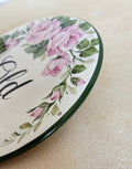 A close-up of the border of a decorative round wall plaque featuring pink roses, green leaves, and black script lettering, shown against a textured beige wall.