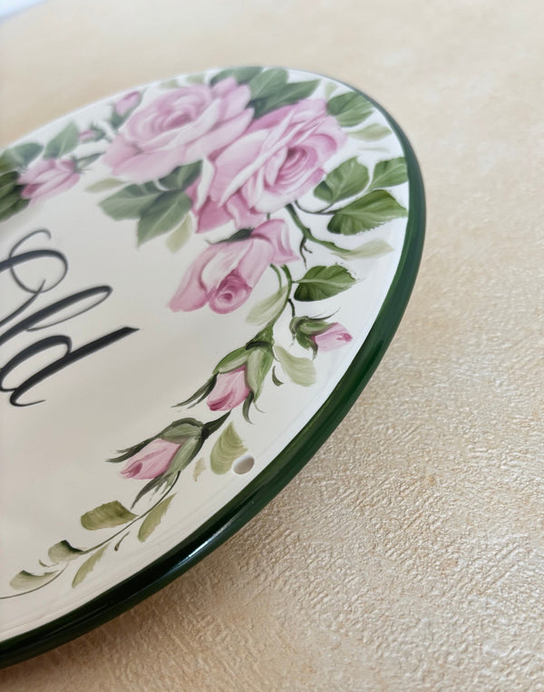 A close-up of the border of a decorative round wall plaque featuring pink roses, green leaves, and black script lettering, shown against a textured beige wall.