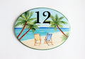 Ceramic house number oval shaped hand painted with a central beach scene featuring two chairs and palm trees, with the number 12 painted on top