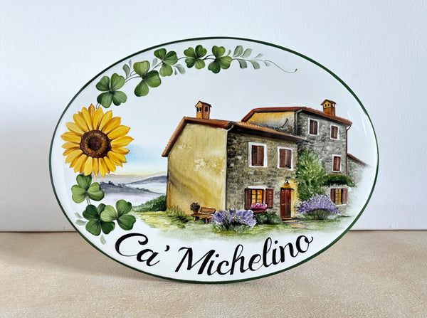 An oval ceramic plaque featuring a hand-painted illustration of a Tuscan stone house, a yellow sunflower, green clover, and the text 'Ca' Michelino' in black script.