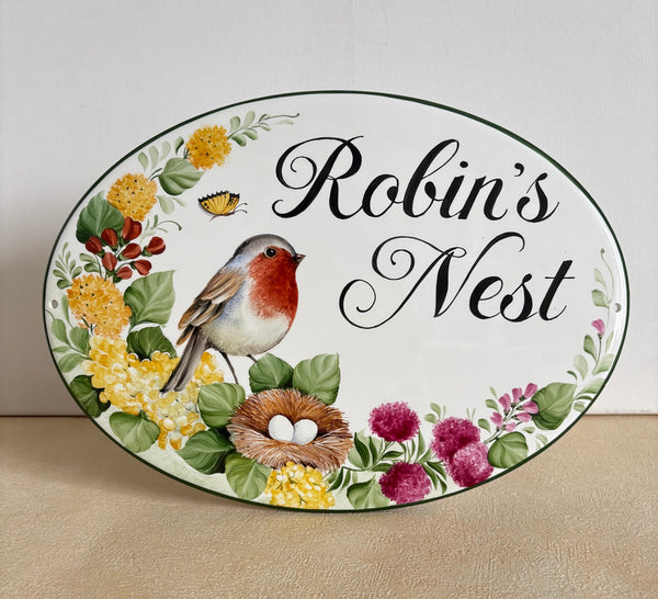 An oval decorative wall House name sign featuring the words 'Robin's Nest' in black script, hand painted with flowers, leaves, a robin bird, and a nest. 
