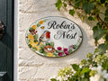 An oval decorative wall sign reading “Robin’s Nest” hangs on a textured cream wall. The sign is hand painted with a robin bird, a nest with white eggs, yellow and pink flowers, green leaves, and a small butterfly.