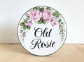 A round decorative ceramic House plaque with a pink roses wreath design, featuring the text 'Old Rosie' in black script