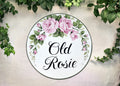 A round white decorative House name sign with a pink rose floral border and the text 'Old Rosie' in black script, displayed against a light textured wall with green ivy around the edges.