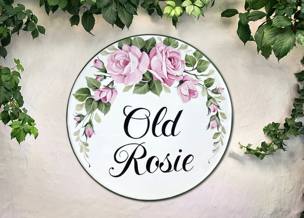 A round white decorative House name sign with a pink rose floral border and the text 'Old Rosie' in black script, displayed against a light textured wall with green ivy around the edges.