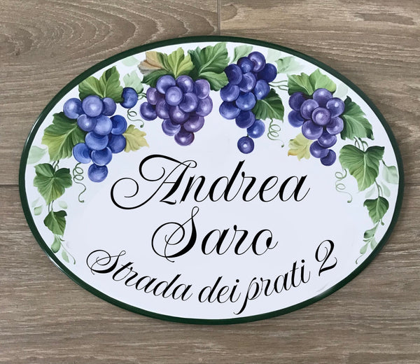 An oval personalized ceramic sign featuring a painted grapevine design with purple grapes and green leaves on a white background, with the names 'Andrea' and 'Saro' and the address 'Strada dei prati 2' written in black script. The sign is photographed flat on a brown wood-grain surface.
