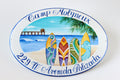 Ceramic house sign for a seaside home, hand painted with colorful surfboards on a beach, the sea, a pier, and a palm branch; the house name is painted at the top and the address at the bottom.