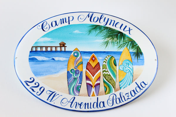 Ceramic house sign for a seaside home, hand painted with colorful surfboards on a beach, the sea, a pier, and a palm branch; the house name is painted at the top and the address at the bottom.