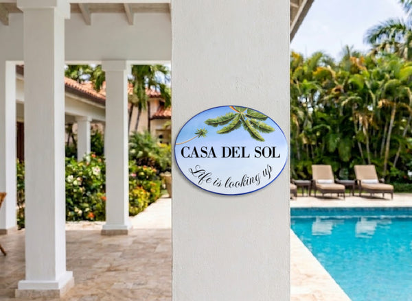 An oval decorative wall sign reading 'CASA DEL SOL' and 'Life is looking up' with green palm leaves, mounted on a white pillar in a resort-like setting with a swimming pool and palm trees in the background.