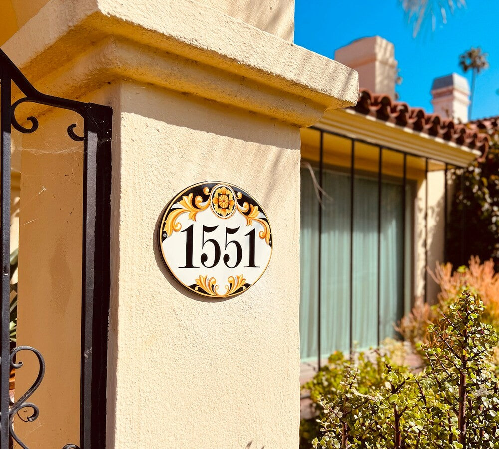 An oval white house number plaque with black numerals '1551' hand painted with gold decorative ornament, mounted on a beige stucco pillar outside a home.