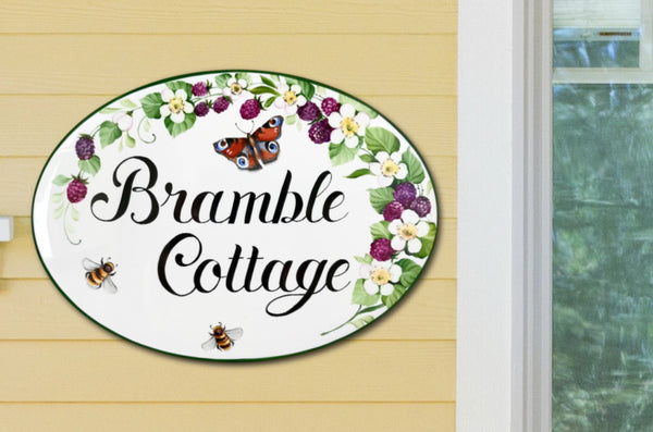 ceramic oval house sign hand-painted with blackberries, white flowers, bees and a red butterfly, mounted on a front door