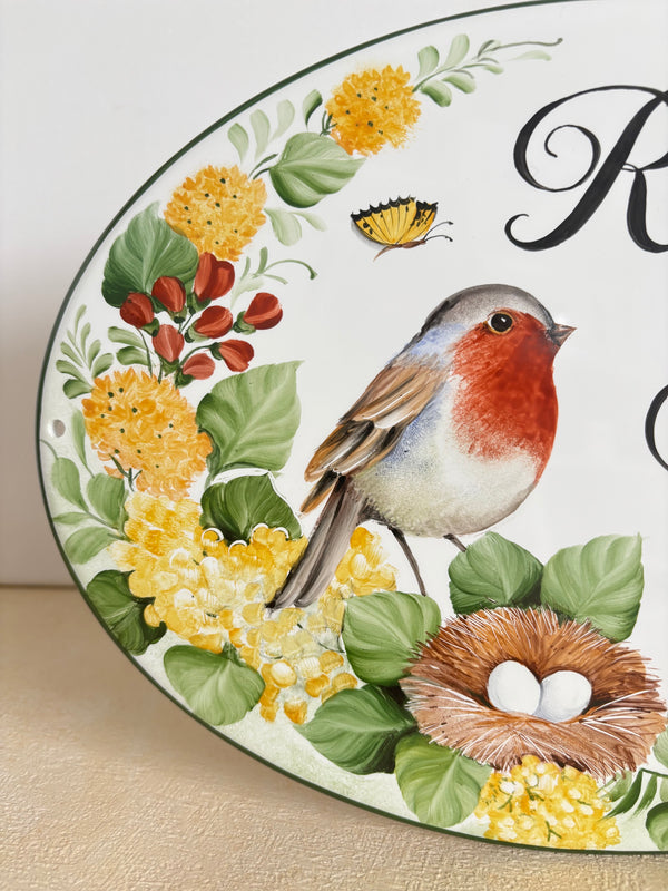 A close-up of a decorative oval wall plaque or sign featuring a painted bird, nest with two white eggs, yellow flowers, green leaves, and a butterfly on a white background with black script lettering.