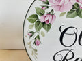 Close-up of a round house sign featuring hand-painted pink roses with green leaves and black script lettering, photographed against a light neutral background.