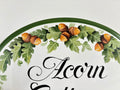 Close-up of a white decorative sign featuring a hand-painted green oak leaf and acorn garland with the word 'Acorn' in black script.
