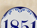 A close-up of a white house number plaque with hand-painted blue deecoration and the numbers 1851 in blue, mounted against a textured beige wall.