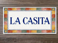 A rectangular ceramic tile sign with the words 'LA CASITA' in blue lettering on a white center panel, surrounded by a multicolor painted border, shown on a dark wood background.