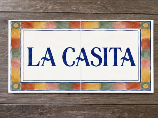 A rectangular ceramic tile sign with the words 'LA CASITA' in blue lettering on a white center panel, surrounded by a multicolor painted border, shown on a dark wood background.