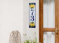 A vertical rectangular ceramic house number plaque with the numbers 7 and 3, hand painted with yellow lemons and green leaves, mounted on a textured white wall next to a wooden door.