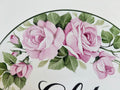 A close-up of a round ceramic plaque featuring painted pink roses with green leaves and part of a black script at the bottom.