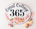 Ceramic house number sign oval shaped hand painted with pink shells and corals on the bottom. The house number is painted in center and the name house is curved on top