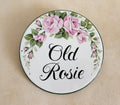 A round ceramic house plaque hand-painted with a floral border of pink roses and green leaves, featuring the text 'Old Rosie' in black script, placed on a beige textured background.