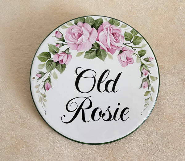 A round ceramic house plaque hand-painted with a floral border of pink roses and green leaves, featuring the text 'Old Rosie' in black script, placed on a beige textured background.