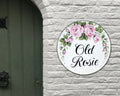 A round white ceramic personalized house sign hand-painted with pink roses and green leaves, featuring the text 'Old Rosie' in black script, mounted on a white brick wall next to a dark green door.