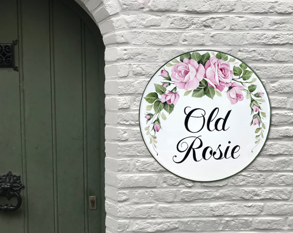 A round white ceramic personalized house sign hand-painted with pink roses and green leaves, featuring the text 'Old Rosie' in black script, mounted on a white brick wall next to a dark green door.