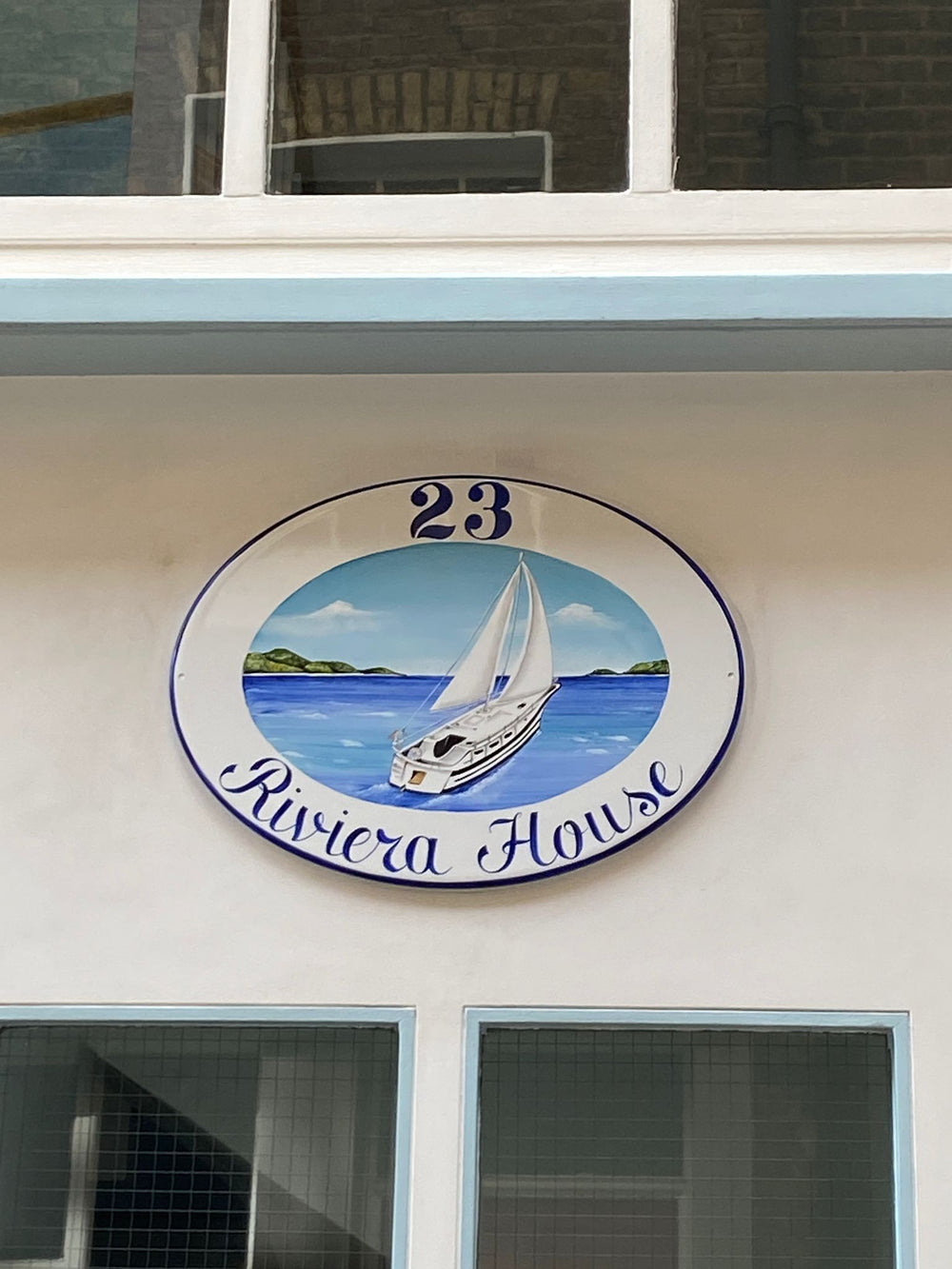 An oval ceramic sign mounted on a white building wall, hand painted with a sailboat on blue water with the text '23 Riviera House'.
