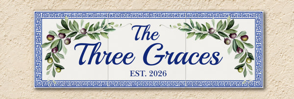 A rectangular decorative wall sign hand painted with a blue Greek key border and watercolor olive branches with  'The Three Graces' and 'EST. 2026' in blue script, shown against a beige textured wall.