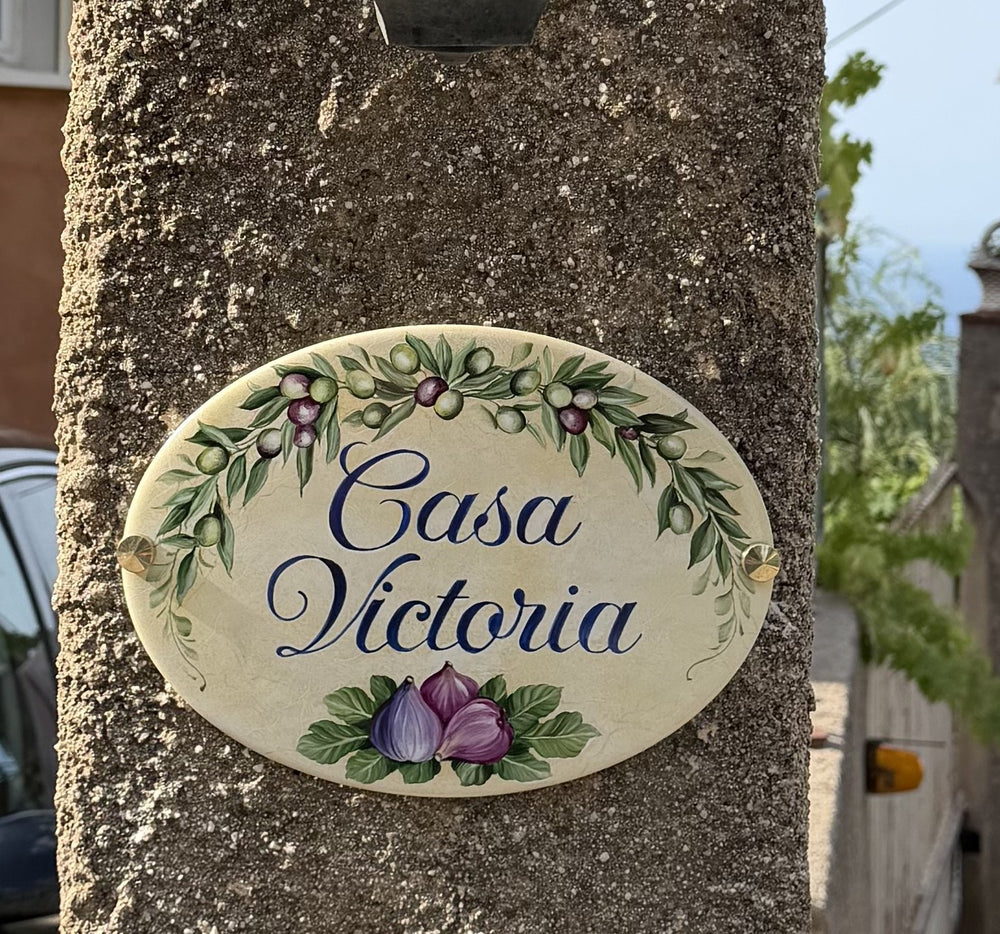An oval ceramic sign reading 'Casa Victoria' in blue script, hand painted with olive branches and figs